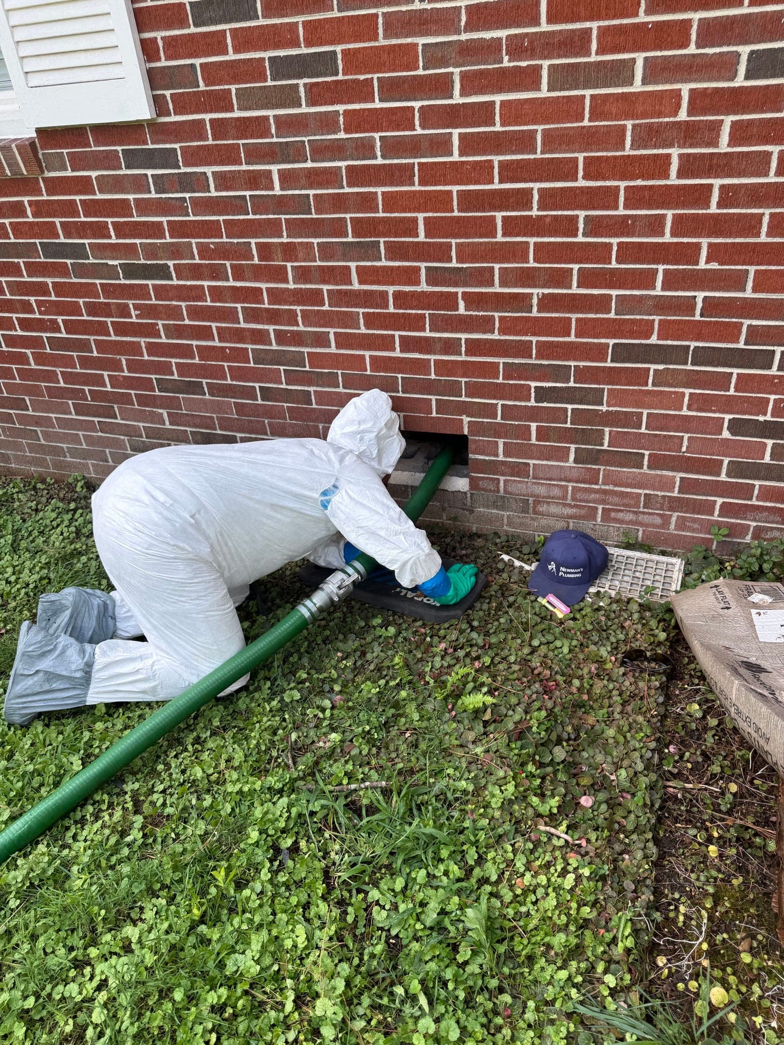 Technician in protective coveralls feeding a suction hose through a crawlspace vent during a sewage cleanup at a Portsmouth VA home — Newman's Plumbing Service & Repair
